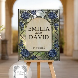 Op de afbeelding: Een rechthoekig glas-in-lood bord met een centraal boogvormig paneel met de tekst EMILIA and DAVID en de datum 05.15.2026. De rand is versierd met blauwe hortensiabloemen en groene bladeren in een geometrisch glas-in-lood frame.