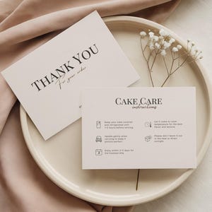 May include: Two white cards with black text on a cream-colored plate. One card reads "THANK YOU For your order" and the other reads "CAKE CARE instructions" with care tips. A sprig of white flowers is placed on the plate.