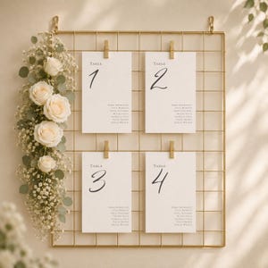 May include: A gold wire grid display featuring four white rectangular cards labelled Table 1, 2, 3, and 4, each held by a gold clip. A vertical arrangement of white roses and small white baby's breath flowers with green leaves is attached to the left side.