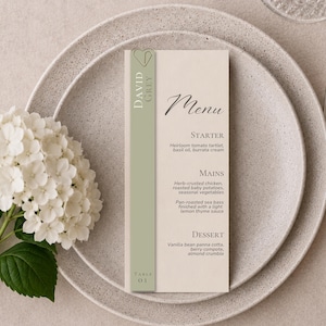 May include: A vertical menu card on a speckled plate with a white hydrangea. The card features a sage green side panel with a heart icon, the text David Grey, and Table 01. The menu lists Starter, Mains, and Dessert with specific food descriptions.