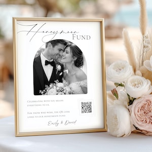 Könnte beinhalten: Ein gerahmtes Schild mit den Worten "Honeymoon Fund" und einem Schwarz-Weiß-Foto eines Paares. Das Schild enthält Text und einen QR-Code, mit den Namen "Emily & David" unten. Ein Strauß weißer und rosa Blumen befindet sich im Hintergrund.