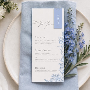 May include: A white rectangular menu card featuring blue floral illustrations along the right edge and the text The Menu, Starter, Main Course, and Dessert. A small blue tag with the name Laura is attached to the top with a silver paperclip.