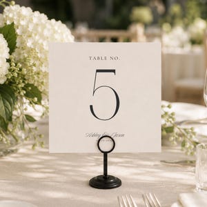May include: A square off-white table number card featuring the text TABLE NO. and the large black numeral 5, with the names Ashley & Jason printed below. The card is held upright by a circular black metal stand on a flat surface.