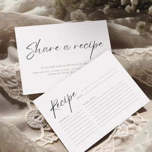 Peut inclure: Deux cartes de recettes blanches avec une écriture noire. L'une des cartes indique "Share a recipe" et l'autre "Recipe". Les cartes sont sur une surface texturée blanc cassé. Les cartes de recettes ont des lignes pour les ingrédients et les instructions.