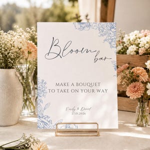May include: A rectangular white sign with blue floral illustrations in the corners, featuring the text "Bloom bar", "MAKE A BOUQUET TO TAKE ON YOUR WAY", and "Emily & David 27.01.2026". The sign is held upright by a thin, gold-toned metal stand.