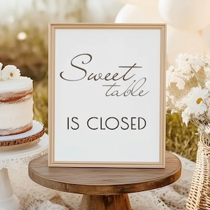 May include: A framed sign with the words "Sweet Table Is Closed" in black script and block letters. A white cake with a floral decoration sits on a white cake stand. A basket of white flowers is also visible.