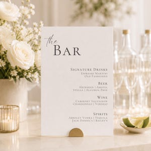 Puede incluir: Un letrero de menú de bar rectangular blanco con texto gris oscuro que dice The Bar, Signature Drinks, Beer, Wine y Spirits, sostenido por un pequeño soporte dorado semicircular. El menú enumera varias opciones de bebidas, incluido Espresso Martini.