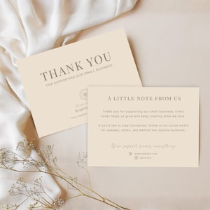Può includere: Due biglietti color crema con testo, uno con la scritta "THANK YOU" e l'altro "A LITTLE NOTE FROM US". I biglietti sono su una superficie beige con un tessuto bianco e un rametto di fiori secchi. Il testo è di colore grigio scuro.