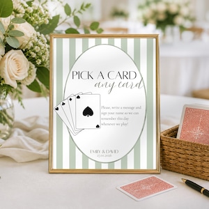 May include: A framed sign with the text "PICK A CARD any card" and a deck of cards illustration. A deck of cards in a woven basket and a pen are next to the sign. The sign has a gold frame and a green and white striped background.