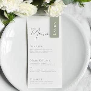 Può includere: Un piatto bianco con un menu e una mise en place. Il menu recita "The Menu" ed elenca un antipasto, un piatto principale e un dessert. Una piccola etichetta verde con il nome "Laura" è attaccata al menu. Fiori bianchi e verde sullo sfondo.