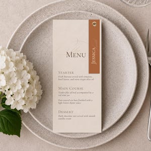 Könnte beinhalten: Eine rechteckige, cremefarbene Menükarte mit dem Text MENU, STARTER, MAIN COURSE und DESSERT sowie einem vertikalen, rostfarbenen Akzentstreifen auf der rechten Seite, der den Namen JESSICA in weißer Schrift und eine kleine goldene Metallöse enthält.