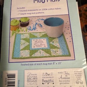 May include: A June Tailor Uplifting Inspirational Mug Mats craft kit package featuring 7 printed statements on 100% cotton fabric and 7 patterns. The packaging shows a finished 8 inch by 10 inch green and blue quilted mug mat with a cup.