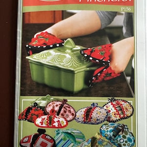 May include: A sewing pattern booklet titled Vanilla House Designs Pot Pinchers! P156. The cover features a green ceramic casserole dish being held by fabric pot pinchers with a red strawberry print and black and white polka dot trim.