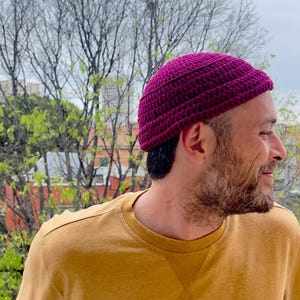 Può includere: Una persona indossa un berretto a maglia di colore viola. Il berretto ha un design semplice con una trama a coste. La persona indossa una maglietta gialla a maniche lunghe con una toppa nera sul lato inferiore destro.