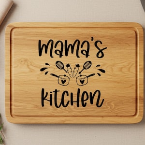 May include: A wooden cutting board with the text "Mama's Kitchen" in black lettering. The text is surrounded by a design of two pots with hearts and carrots.
