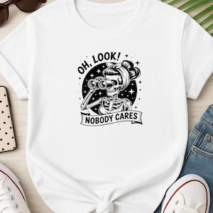 May include: White t-shirt featuring a black and white graphic of a skeleton with binoculars, a headscarf, and the text "OH, LOOK!" and "NOBODY CARES". The shirt is paired with blue jeans and white sneakers.