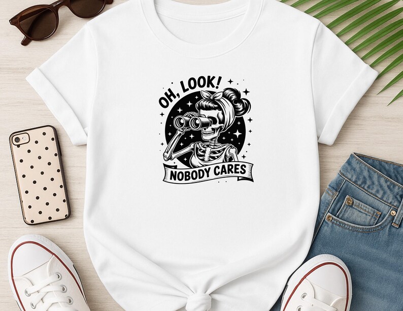 May include: White t-shirt featuring a black and white graphic of a skeleton with binoculars, a headscarf, and the text "OH, LOOK!" and "NOBODY CARES". The shirt is paired with blue jeans and white sneakers.