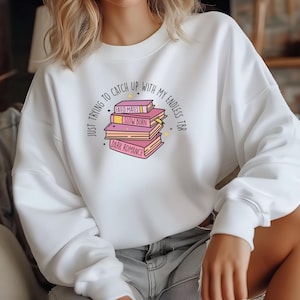 May include: White sweatshirt with a graphic of a stack of books with the text "JUST TRYING TO CATCH UP WITH MY ENDLESS TBR" and the titles of the books "FATED MATES", "SLOW BURN", and "DARK ROMANCE".