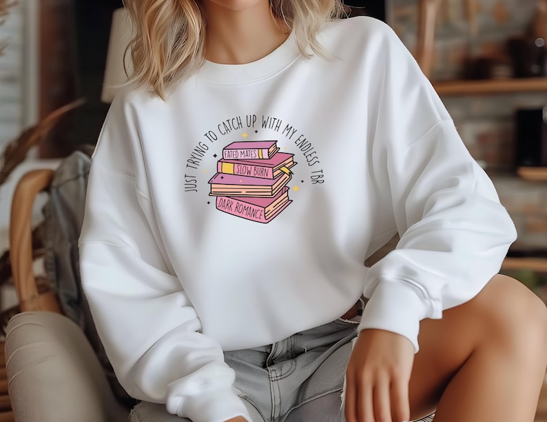 May include: White sweatshirt with a graphic of a stack of books with the text "JUST TRYING TO CATCH UP WITH MY ENDLESS TBR" and the titles of the books "FATED MATES", "SLOW BURN", and "DARK ROMANCE".