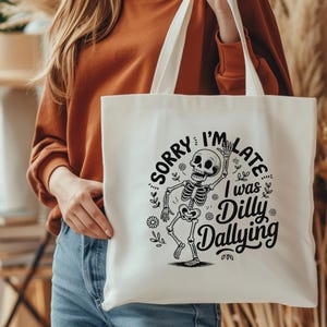 May include: Cream-colored tote bag with a black skeleton graphic and the text "SORRY I'M LATE I WAS DILLY DALLYING". The bag has two white handles and is held by a person.
