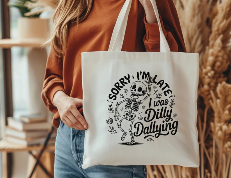 May include: Cream-colored tote bag with a black skeleton graphic and the text "SORRY I'M LATE I WAS DILLY DALLYING". The bag has two white handles and is held by a person.