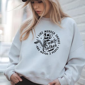 May include: White sweatshirt with a black graphic of a skeleton and the text "I like murder shows, comfy clothes, and maybe 3 people." The design is in a circular format. The sweatshirt is a casual style.