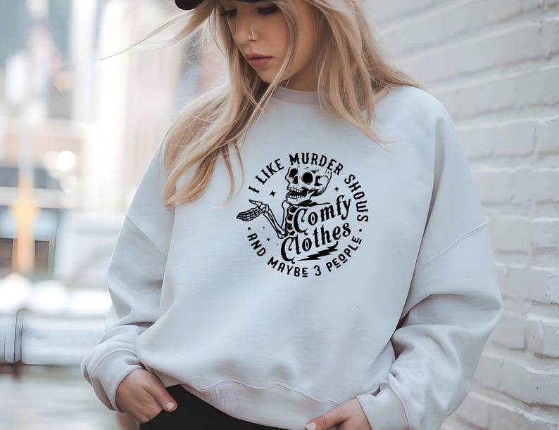 May include: White sweatshirt with a black graphic of a skeleton and the text "I like murder shows, comfy clothes, and maybe 3 people." The design is in a circular format. The sweatshirt is a casual style.