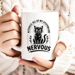 May include: White ceramic mug featuring a black cat illustration with the text "ACTUALLY, ALL OF MY SYSTEMS ARE NERVOUS". The cat has wide eyes and a nervous expression. The mug has a curved handle.
