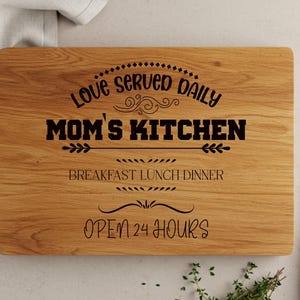 May include: A wooden cutting board with a handle, engraved with the text "Love served daily Mom's Kitchen Breakfast Lunch Dinner Open 24 Hours".