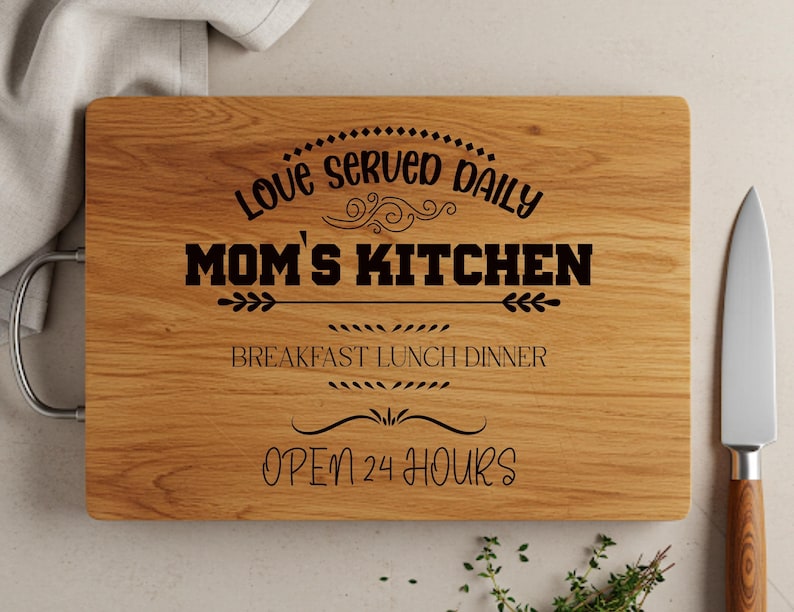 May include: A wooden cutting board with a handle, engraved with the text "Love served daily Mom's Kitchen Breakfast Lunch Dinner Open 24 Hours".