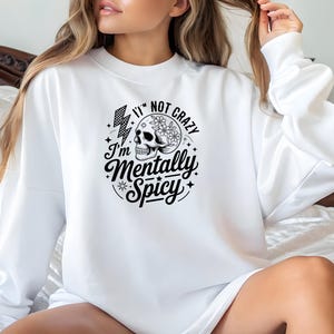 May include: White sweatshirt with a black graphic design. The design includes a skull with floral accents, a lightning bolt, and the text "I'm not crazy, I'm Mentally Spicy" in a circular arrangement. The sweatshirt has long sleeves.