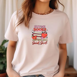May include: Pink t-shirt with a graphic design that says "Buy Me Books and Call Me a Good Girl". The graphic features a stack of books, a heart, and a chili pepper.