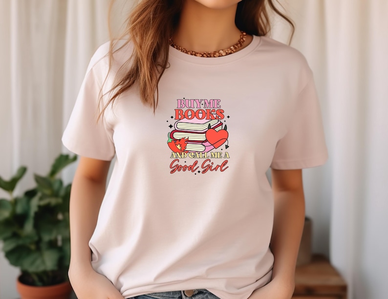 May include: Pink t-shirt with a graphic design that says "Buy Me Books and Call Me a Good Girl". The graphic features a stack of books, a heart, and a chili pepper.