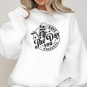 May include: White sweatshirt with a black graphic of a skeleton inside a circle. The text reads "Have The Day You Deserve". The skeleton is holding a hand out. The sweatshirt has long sleeves and a crew neck.