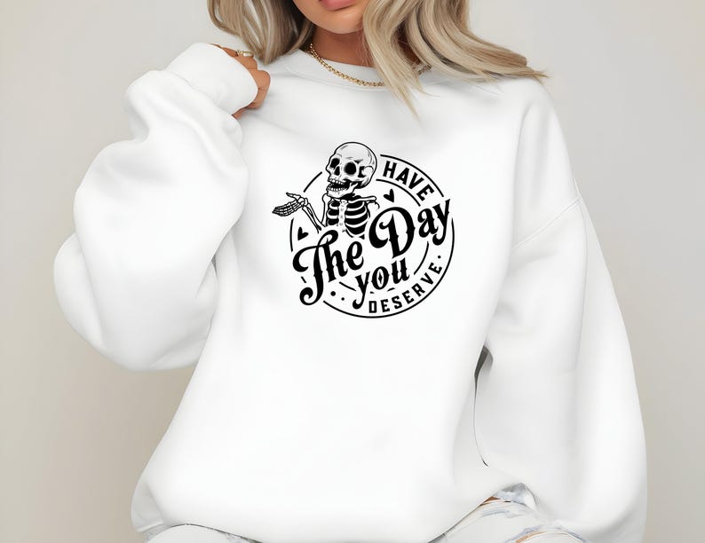 May include: White sweatshirt with a black graphic of a skeleton inside a circle. The text reads "Have The Day You Deserve". The skeleton is holding a hand out. The sweatshirt has long sleeves and a crew neck.