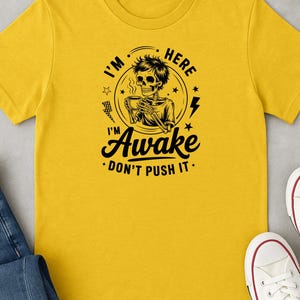 May include: Yellow t-shirt with a black graphic of a skeleton holding a coffee cup, with the text "I'm Here Awake Don't Push It." The shirt is paired with blue jeans and white sneakers.