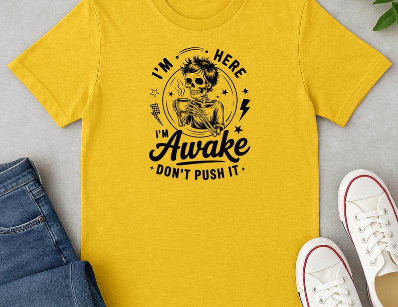May include: Yellow t-shirt with a black graphic of a skeleton holding a coffee cup, with the text "I'm Here Awake Don't Push It." The shirt is paired with blue jeans and white sneakers.