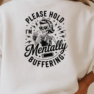 May include: White sweatshirt with a black graphic on the back. The graphic features a skeleton wearing sunglasses and a headscarf, holding a drink. Text reads "PLEASE HOLD I'M Mentally BUFFERING" with a low battery icon.