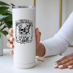 May include: White insulated tumbler with a black graphic of a skull with a butterfly and rose, and the text "I believe in holding grudges, I'll heat in Hell". The tumbler has a silver lid.