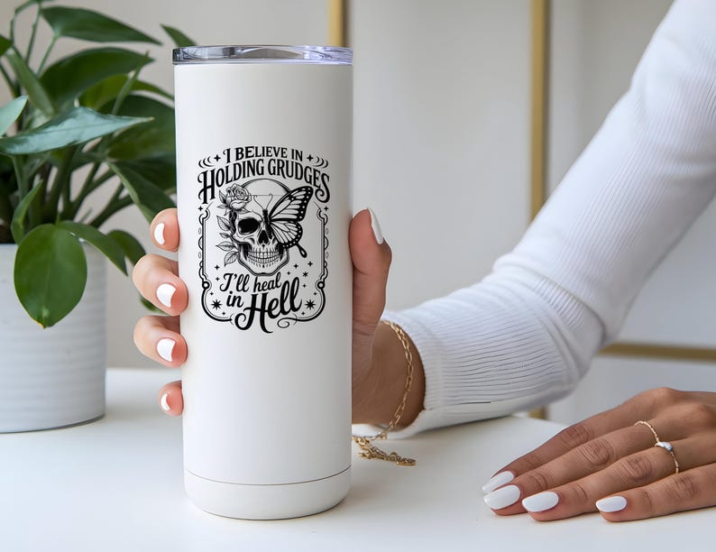 May include: White insulated tumbler with a black graphic of a skull with a butterfly and rose, and the text "I believe in holding grudges, I'll heat in Hell". The tumbler has a silver lid.