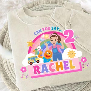 Puede incluir: Sudadera beige con un diseño gráfico colorido para una celebración de cumpleaños. El diseño incluye un arcoíris, personajes de dibujos animados, el número 2 y el nombre "RACHEL" en letras grandes. También está presente el texto "CAN YOU SAY...".