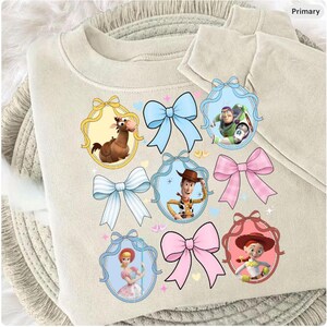 May include: A beige sweatshirt featuring cartoon character portraits within decorative frames and colorful bows. The characters include a horse, Buzz Lightyear, Woody, Bo Peep, and Jessie. The design is playful and whimsical.