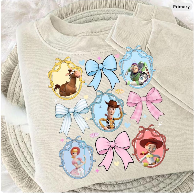 May include: A beige sweatshirt featuring cartoon character portraits within decorative frames and colorful bows. The characters include a horse, Buzz Lightyear, Woody, Bo Peep, and Jessie. The design is playful and whimsical.