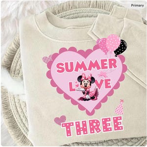 May include: A cream-coloured sweatshirt with a pink heart design. The heart features the words "SUMMER LOVE" and a Minnie Mouse graphic. The word "THREE" is below. Pink and black polka dot balloons and a party hat are also visible.