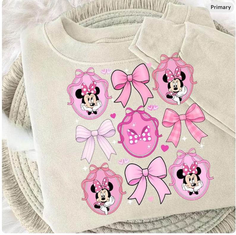 May include: A beige sweatshirt with a design of pink bows and Minnie Mouse in oval frames. The pattern includes different bow styles and small pink hearts, giving a playful and feminine look.
