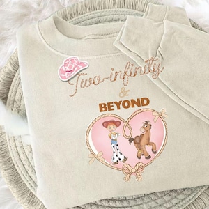 Peut inclure: Un sweat-shirt beige avec le texte "Two-infinity & BEYOND" en &eacute;criture marron. Un autocollant de chapeau de cow-boy rose est en haut &agrave; gauche. Un motif en forme de c&oelig;ur repr&eacute;sente une cowgirl et un cheval de dessin anim&eacute;.