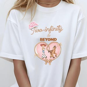 Peut inclure: T-shirt blanc avec un autocollant de chapeau de cow-boy rose. Le t-shirt pr&eacute;sente un motif en forme de c&oelig;ur avec une cowgirl et un cheval, entour&eacute; d'une bordure en corde. Le texte "Two-infinity & BEYOND" est &eacute;crit dans une police d&eacute;corative.