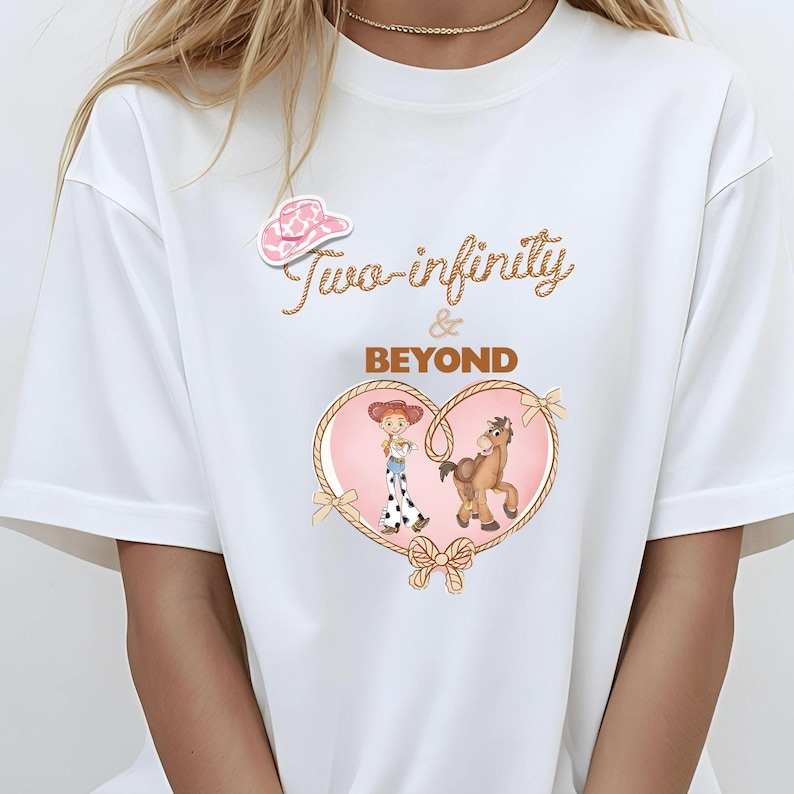 Peut inclure: T-shirt blanc avec un autocollant de chapeau de cow-boy rose. Le t-shirt pr&eacute;sente un motif en forme de c&oelig;ur avec une cowgirl et un cheval, entour&eacute; d'une bordure en corde. Le texte "Two-infinity & BEYOND" est &eacute;crit dans une police d&eacute;corative.