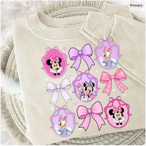 May include: A beige sweatshirt with cartoon graphics of Minnie Mouse and Daisy Duck in pink and purple oval frames, and bows. The design incorporates pink, purple, and white elements.