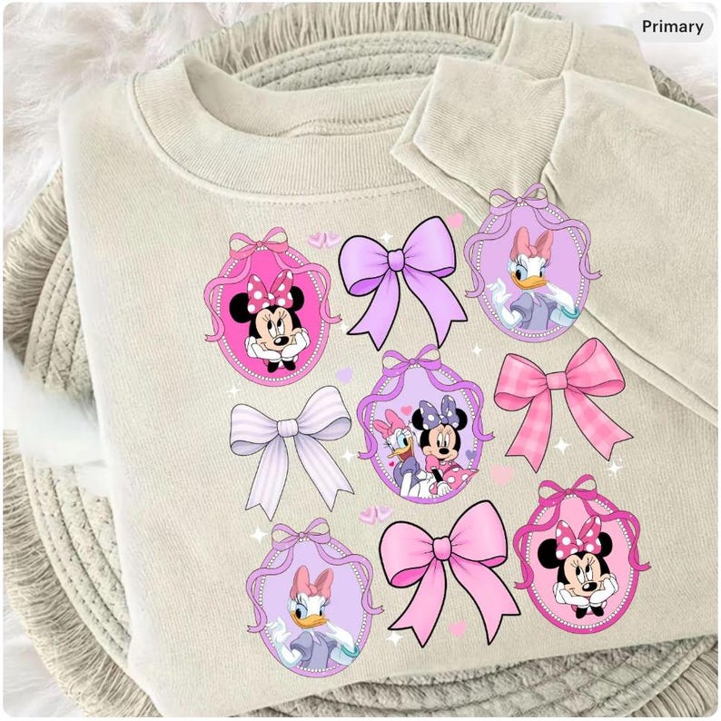 May include: A beige sweatshirt with cartoon graphics of Minnie Mouse and Daisy Duck in pink and purple oval frames, and bows. The design incorporates pink, purple, and white elements.